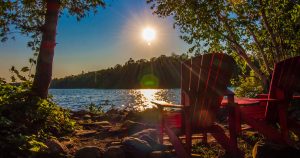 Two red chairs sitting at the edge of a lake with the sun in the sky.