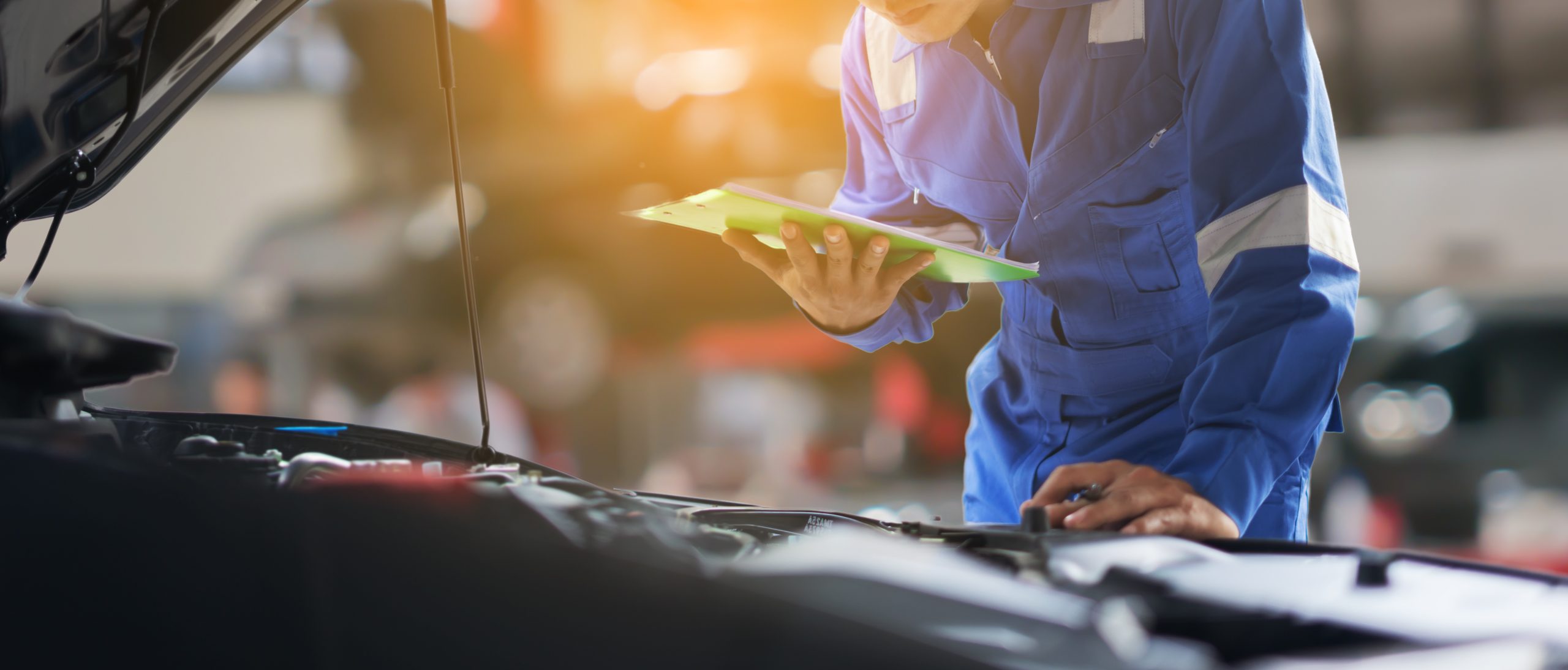 A mechanic inspecting a car engine while holding a clipboard in a workshop.